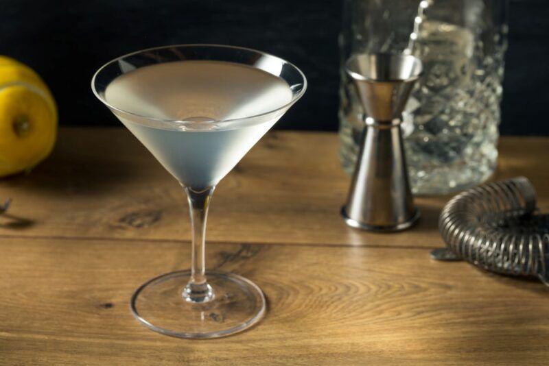 A martini glass on a wooden table next to a cocktail jigger, some spirit, a strainer, and a lemon. The glass contains a spicy shine tini cocktail