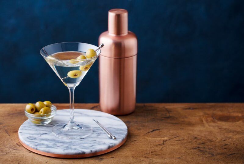 A martini with olives on a tray, next to a small dish of olives and a cocktail shaker