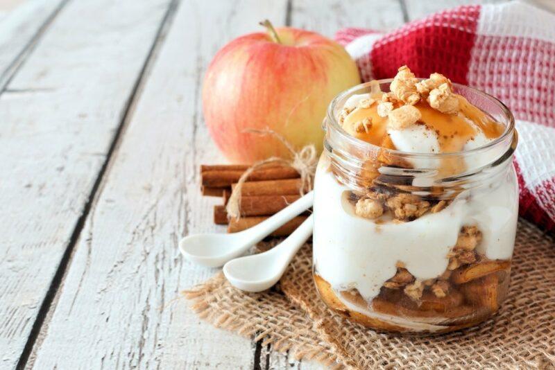 A wooden table with an apple, some cinnamon sticks, two white spoons, and a mason jar with baked apple parfait