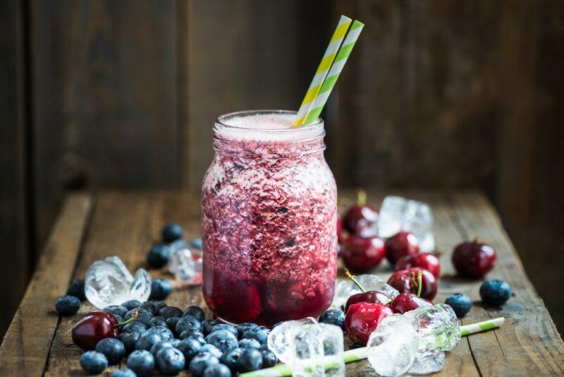 A mason jar filled with crushed berries and two straws. It is surrounded by blueberries, cherries, and plenty of ice