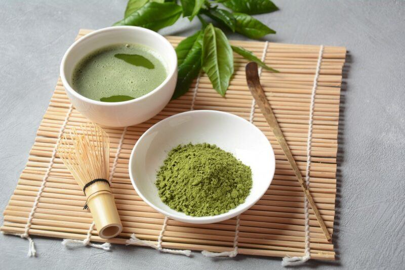 A small wooden bowl of matcha powder, with a small dish of matcha tea in the background, on a bamboo mat next to a whisk