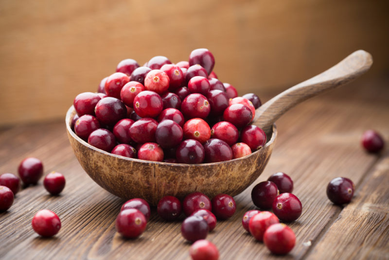 A wooden table with various cranberries scattered around and a metal spoon that is stacked full of cranberries