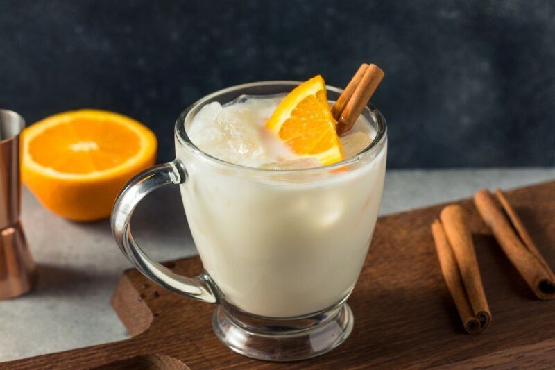 A glass mug containing a milk and honey cocktail that is garnished with a lemon wedge and a stick of cinnamon