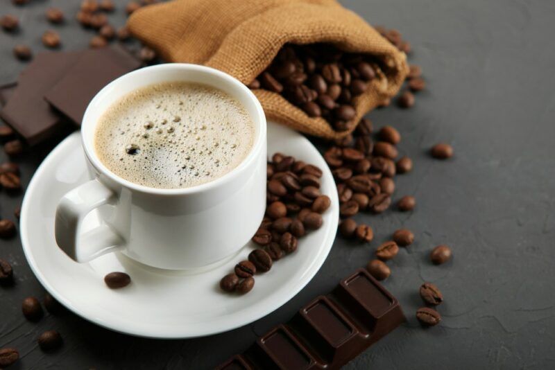 A gray table with a mug of coffee on a sauce, next to a bag of coffee beans that has spilled onto the table and some chocolate