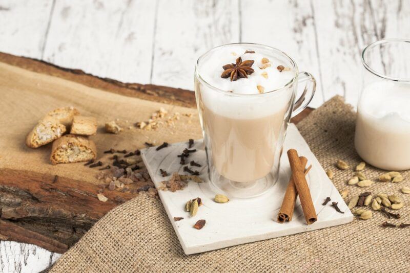 A glass mug of a chai latte on a white wooden table next to a piece of wood