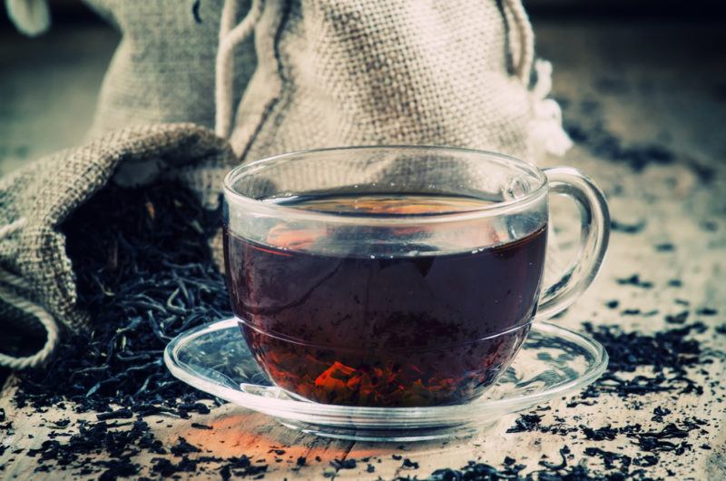 A glass mug of hot black tea with black tea leaves scattered on the table