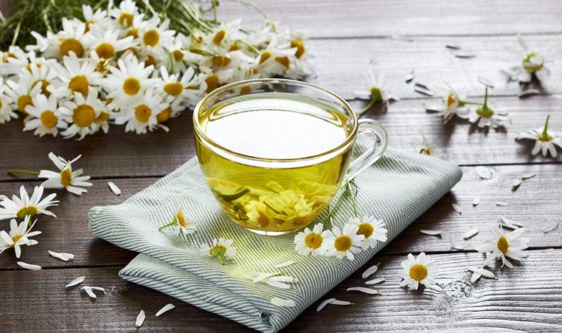 A glass mug of chamomile tea on a cloth mat, surrounded by chamomile flowers