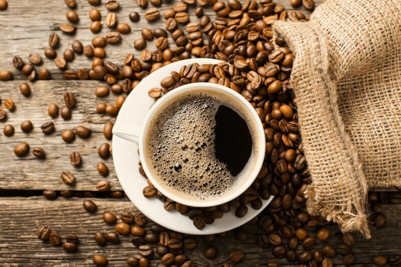 A wooden table with coffee beans strewn across it, with a burlap sack and a mug of hot coffee