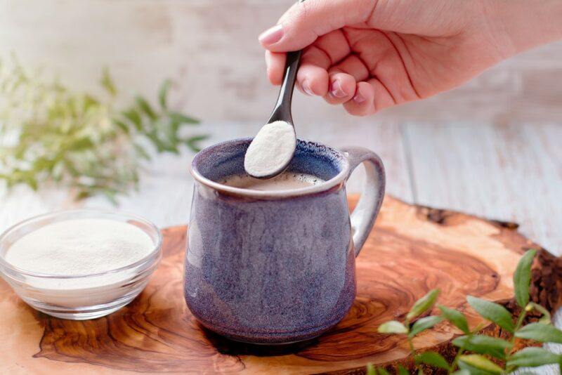 A blue gray mug of coffee that someone is putting a spoonful of collagen powder into. There is a clear bowl of collagen powder next to the coffee