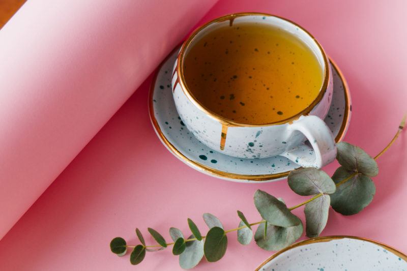 A mug of eucalyptus tea, next to a sprig of the tree on a pink background