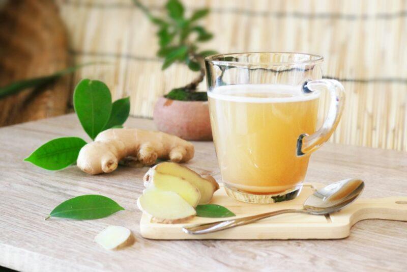 A glass mug of hot homemade ginger tea, next to ginger, some leaves, and a spoon