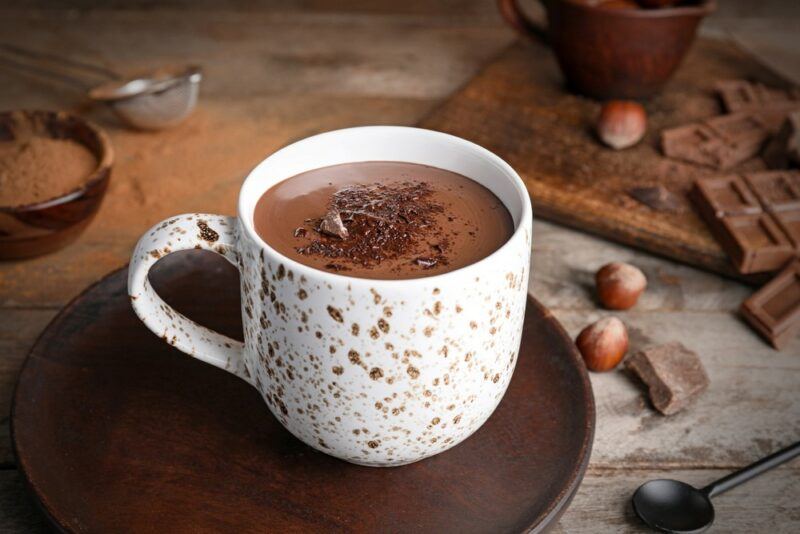 A white mug containing hot chocolate with ingredients in the background