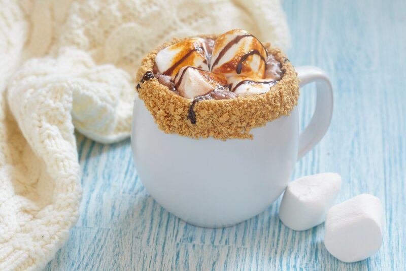 A white mug filled with hot chocolate, with graham crackers around the rim and marshmallows inside