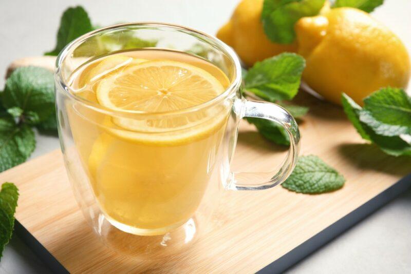 A wooden board with a mug of hot lemon water, with lemons and leaves in the background