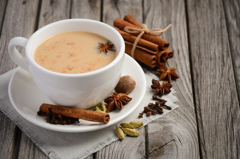 A gray wooden table with a white mug and saucer of masala chai, with a collection of different spices