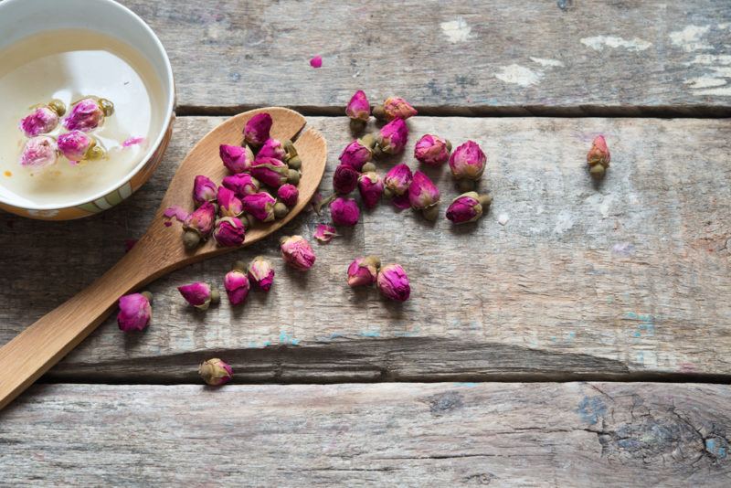 A wooden table with rosebuds, a wooden spoon and a mug of rose tea