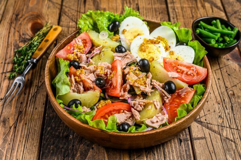 A wooden bowl containing niçoise salad with a knife and a small bowl in the background