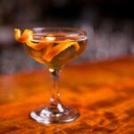 A wooden table with a single nikolaschaka cocktail that's been garnished with a twist of orange