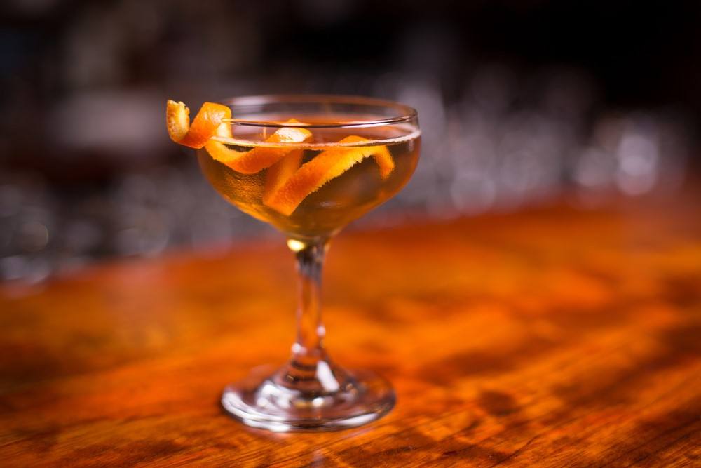 A wooden table with a single nikolaschaka cocktail that's been garnished with a twist of orange