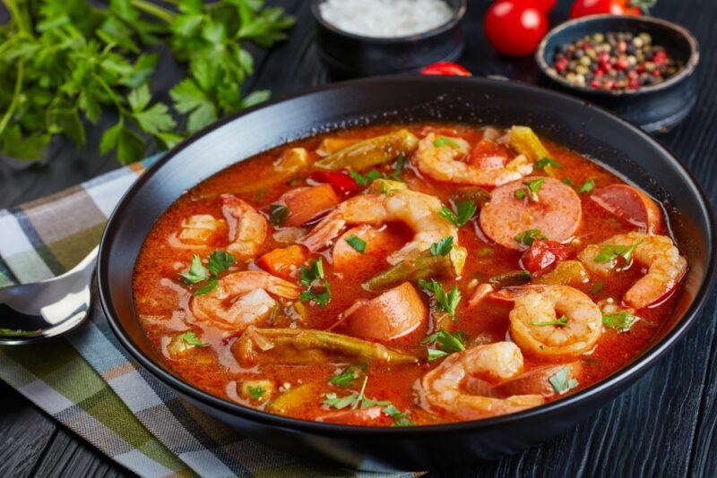 A large black pan containing gumbo with shrimp and meat, in front of fresh ingredients