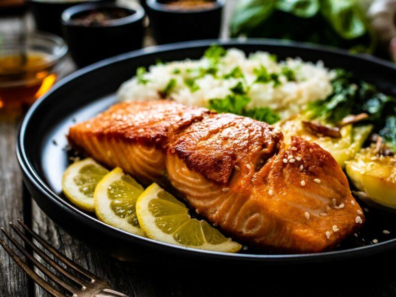 A large black plate with a piece of cooked salmon, potato or rice as a side, and sliced lemons