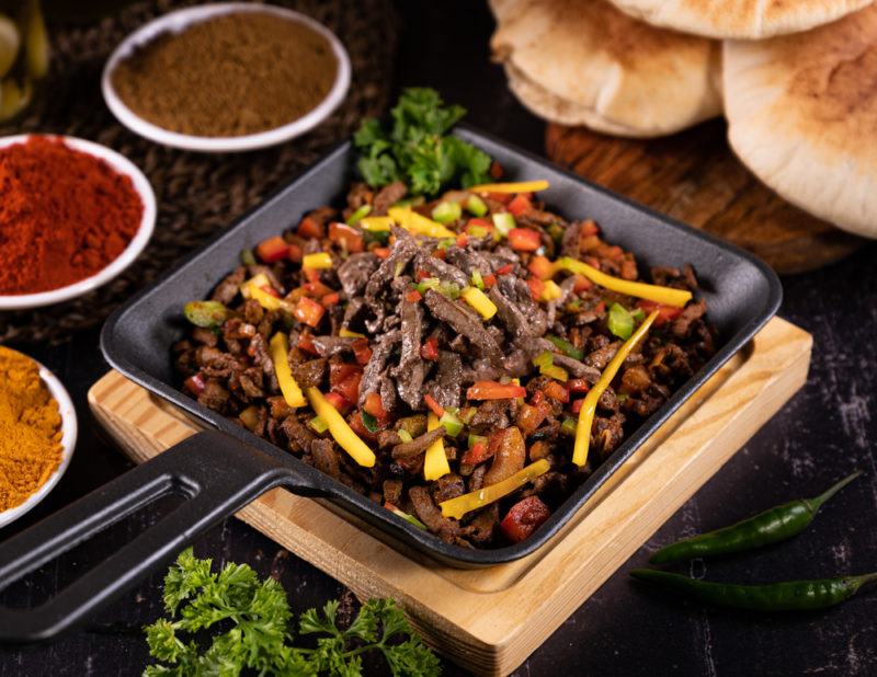 A cast iron fry pan filled with pan seared liver, red and yellow peppers, along with various dips and pieces of bread