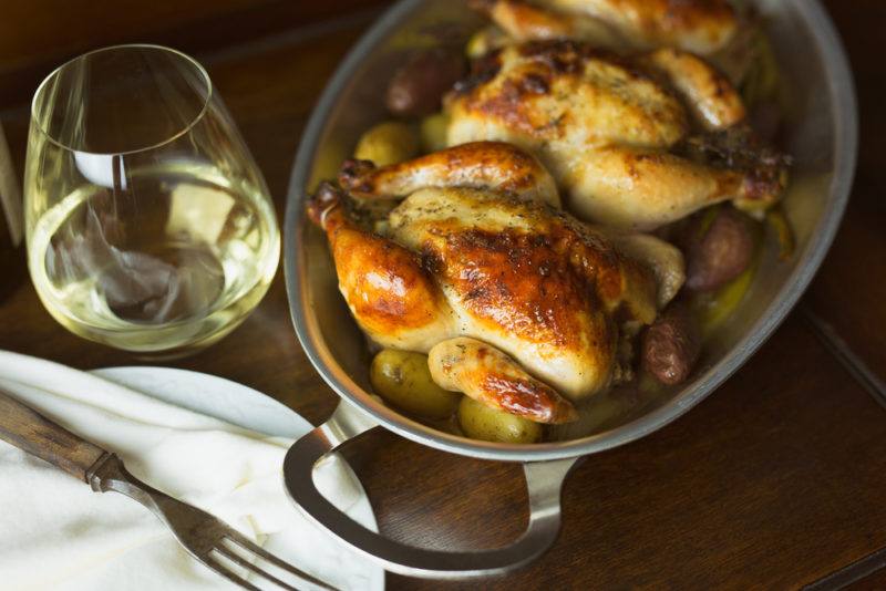 An oven dish with rosted chickens next to a glass of white wine