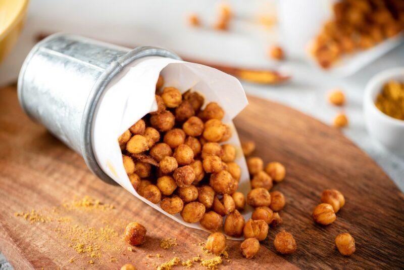 A picnic container of roasted chickpeas that are spilling out onto a wooden board