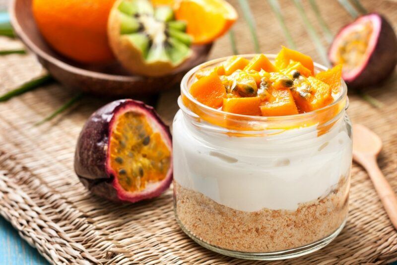 A glass containing a passionfriend cheesecake, next to half a passionfruit and other fruits in a bowl