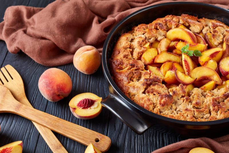 A black dish containing a peach cobbler next to some peaches and wooden serving tools