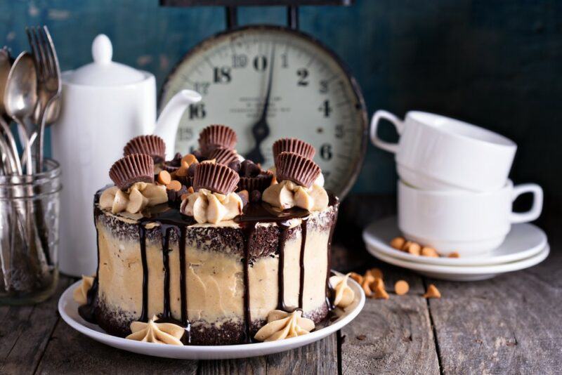 A wooden table with a clock in the background, cups, and a jug, plus a decadent peanut butter and chocolate cake