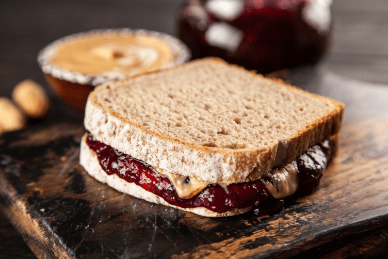 A single peanut butter and jelly sandwich made on brown bread, with a dish of peanut butter in the background.