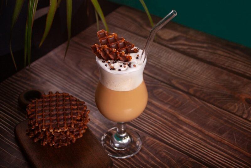 A single glass containing a peanut butter fizz cocktail on a wooden table. There is a stack of chocolate waffles on the table with an extra one garnishing the drink.