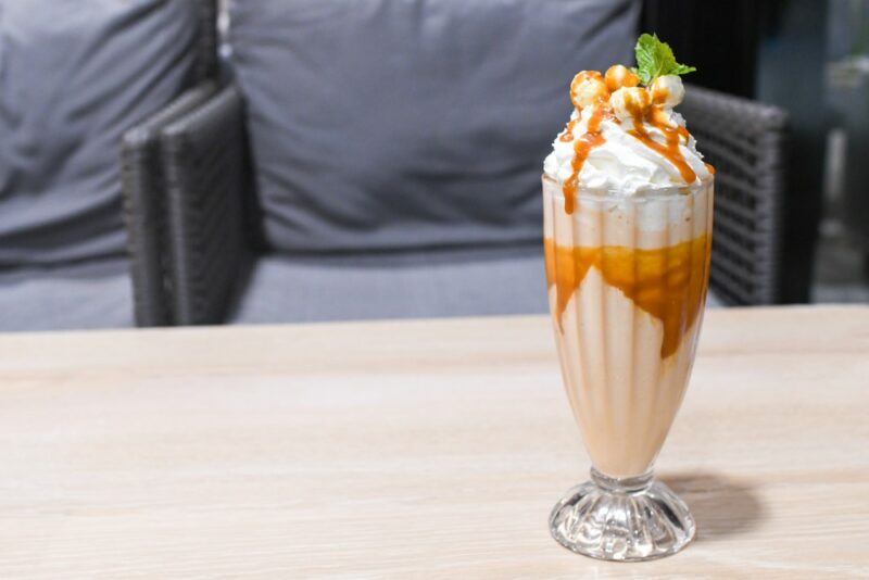 A tall shake glass containing a peanut butter whiskey milkshake, on a table, in front of two chairs