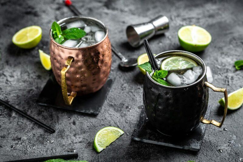 Two Moscow mule cocktails on a gray table with plenty of ice, garnished with mint leaves, with a cocktail jigger and limes on the table