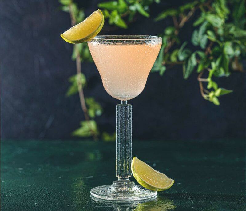 A glass containing a pegu club cocktail on a table with some leaves in the background