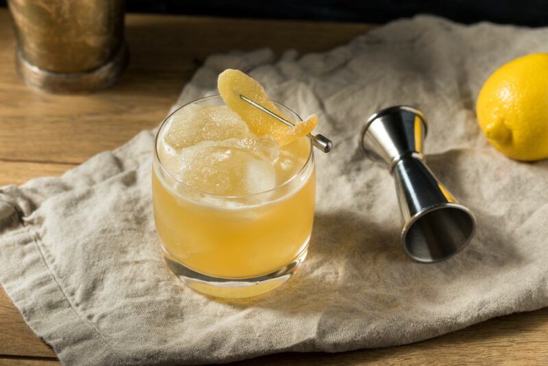 A cloth on a wooden table with a lemon, a cocktail jigger, and a glass containing a penicillin cocktail