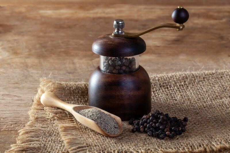 A wooden pepper grinder with freshly ground pepper and some peppercorns in front of it