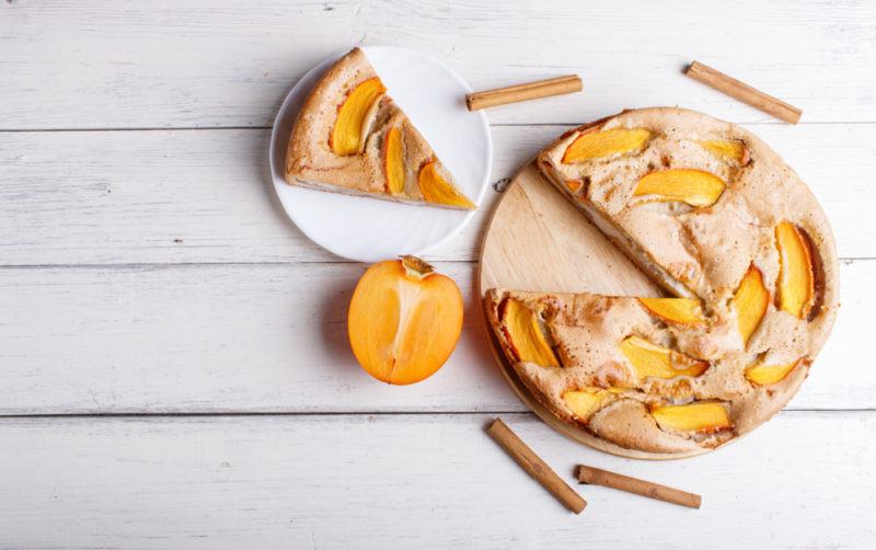 A persimmon pie on a pie dish with one piece cut out that is next to it on a white plate. There are cinnamon sticks scattered around and half of a persimmon