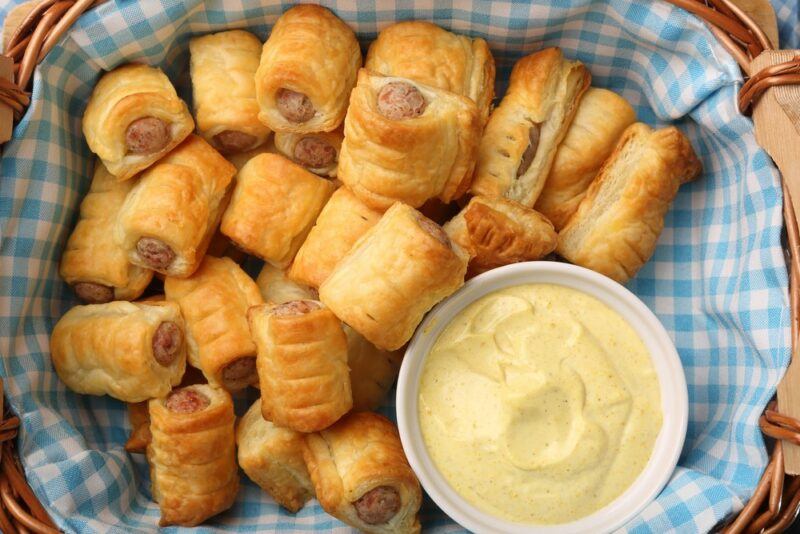 A picnic basket with a blue and white cloth, filled with sausage rolls and a large bowl of dip