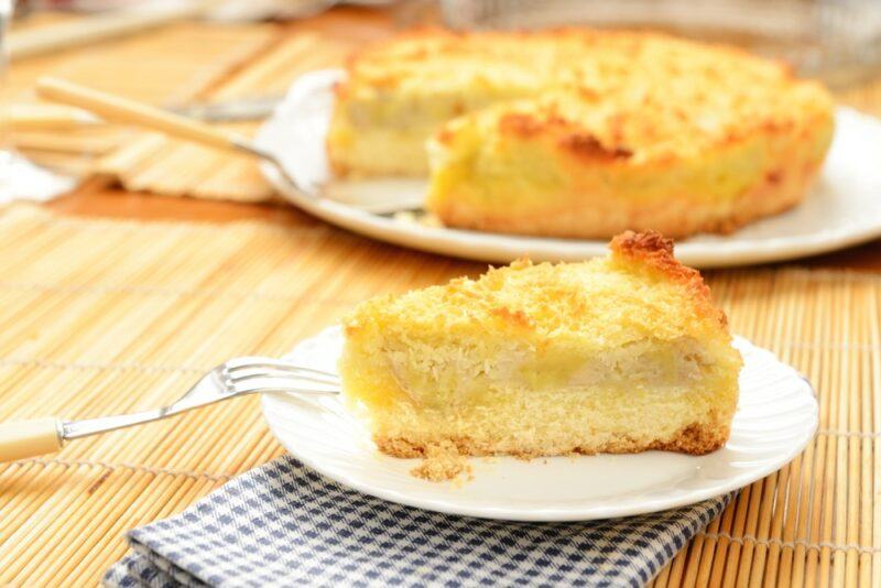 A small white plate with a piece of banana and pineapple cake, with the rest of the cake in the background