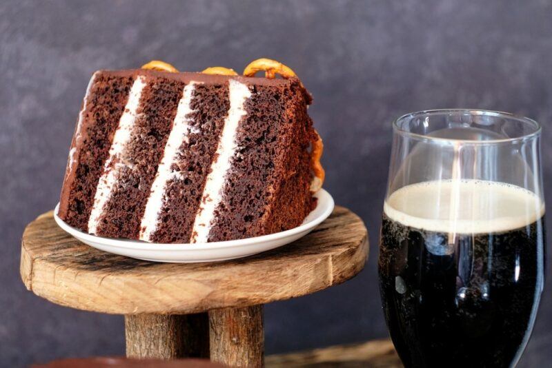 A wooden stand with a large piece of chocolate pretzel cake, next to a mug of chocolate stout