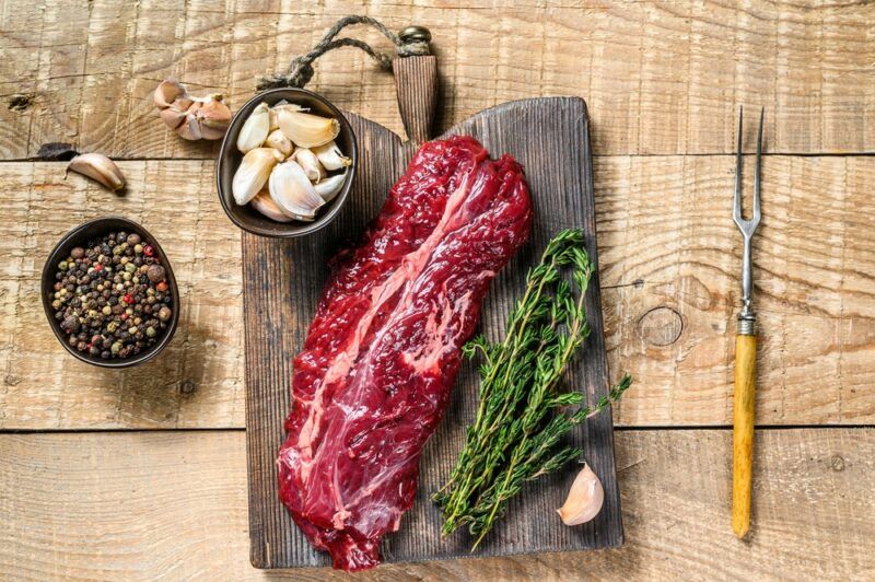 A light wooden table with a wooden board that contains a raw piece of hanger steak, fresh rosemary, a clove of garlic, and a small bowl of garlic cloves