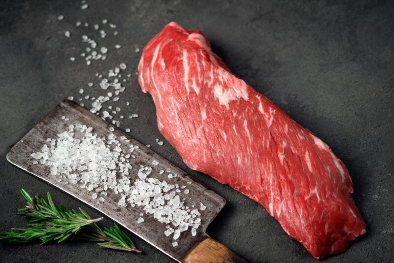 A black table with a raw piece of skirt steak next to a dish containing plenty of salt. There are also a few sprigs of rosemary on the table