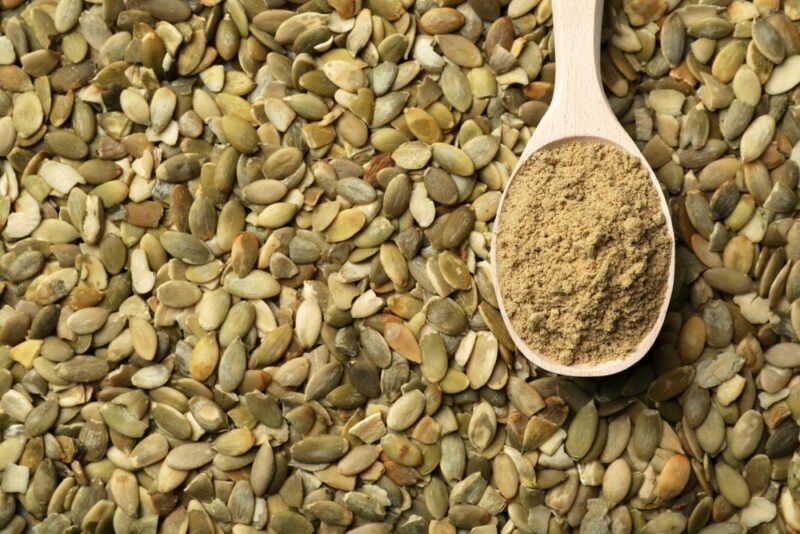 A large pile of pumpkin seeds, with a wooden spoon containing pumpkin seed powder on top