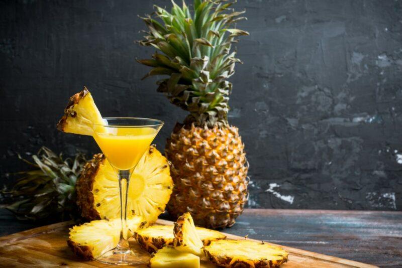 A pineapple martini on a wooden board surrounded by pineapple pieces with a full pineapple in the background