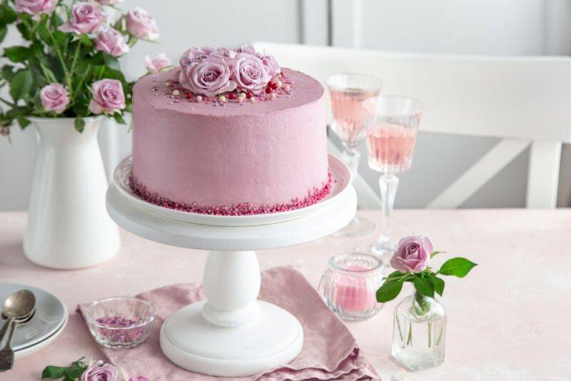 A white cake stand with a pink ricotta and beet cake, next to some pink roses