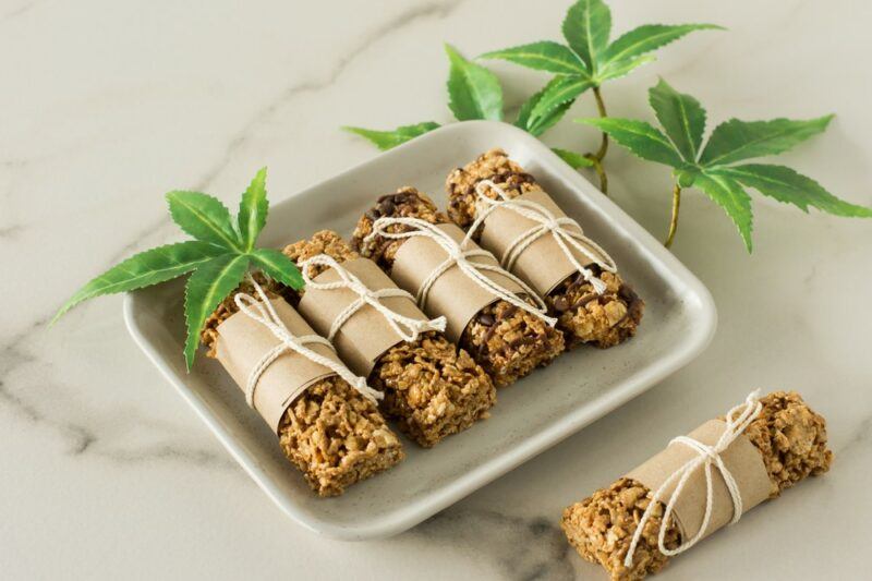 A white plate containing four protein bars wrapped with paper and string, with a fifth on the marble table. There are also some hemp leaves.