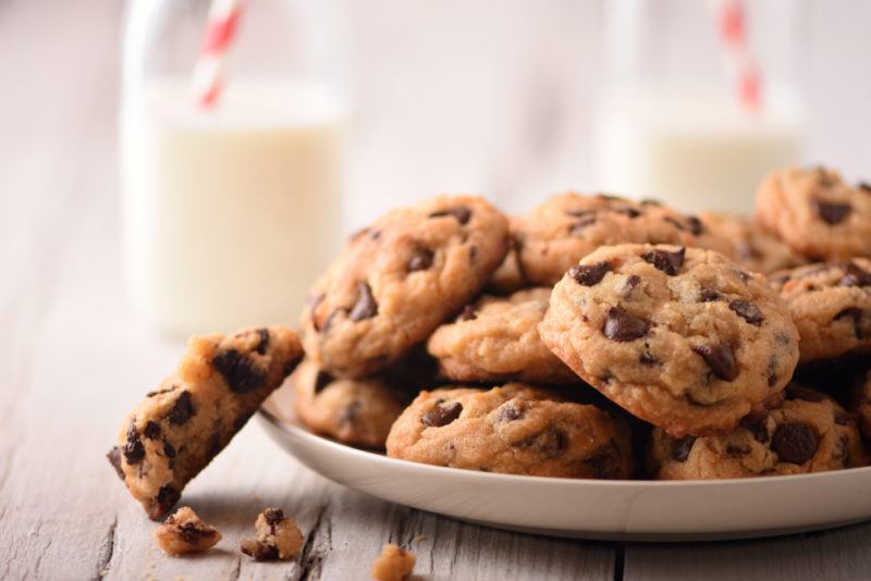 A white dish with chocolate chip cookies and two out-of-focus glasses of milk in the background