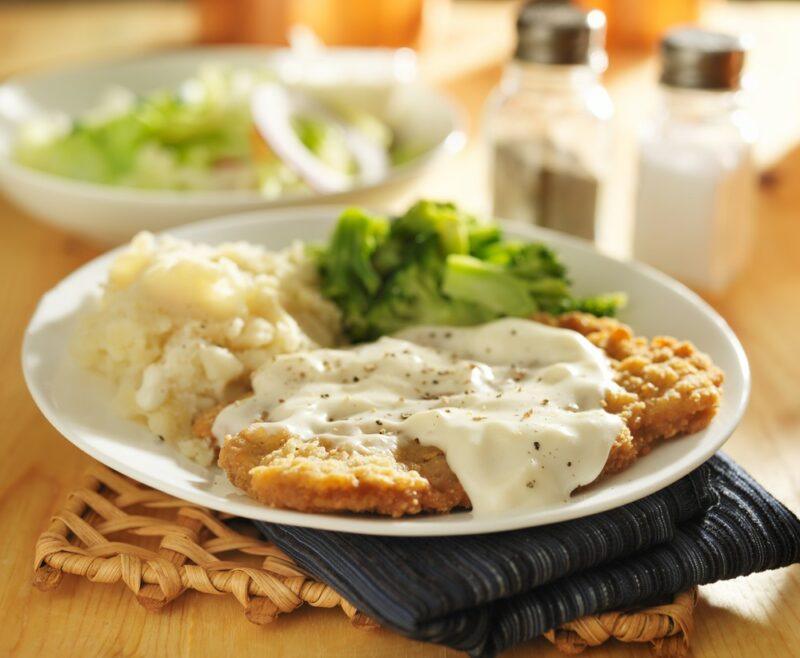 A plate of chicken fried steak or country fried steak with white gravy and broccoli, with a second plate in the background, plus some salt and pepper